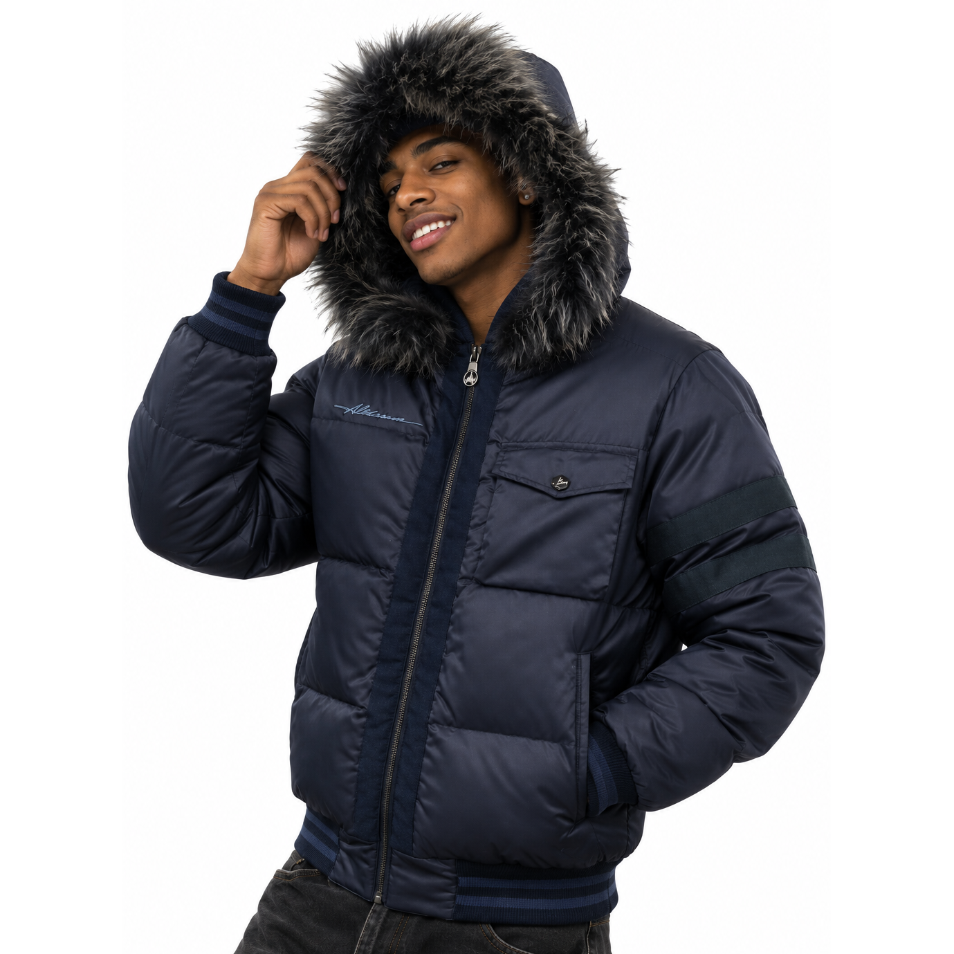 A man wearing a navy blue puffer jacket with a dark gray faux-fur trimmed hood, dark navy striped sleeve detail, and a front zipper, paired with dark jeans against a white background.