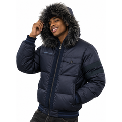A man wearing a navy blue puffer jacket with a dark gray faux-fur trimmed hood, dark navy striped sleeve detail, and a front zipper, paired with dark jeans against a white background.